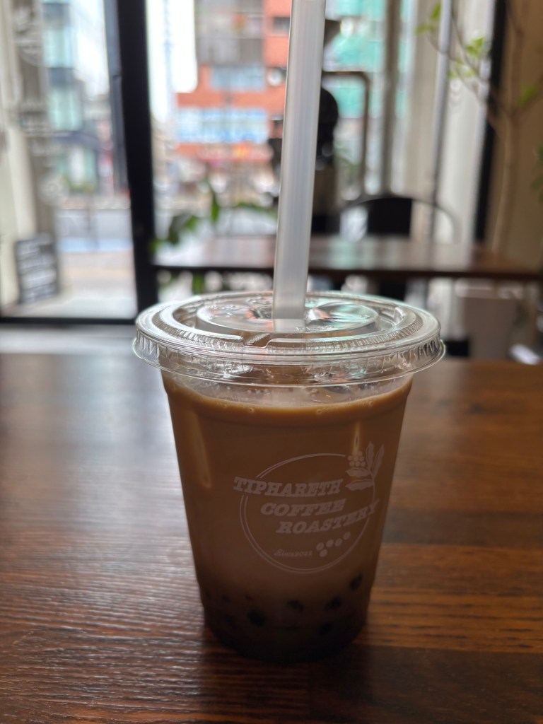 Iced coffee with boba served in a clear cup at Tiphareth Coffee Roastery, with a view of the cafe interior and cityscape in the background.