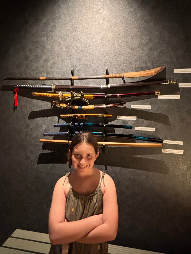 A young child stands confidently with arms crossed in front of a display featuring several traditional Japanese swords mounted on a wall.