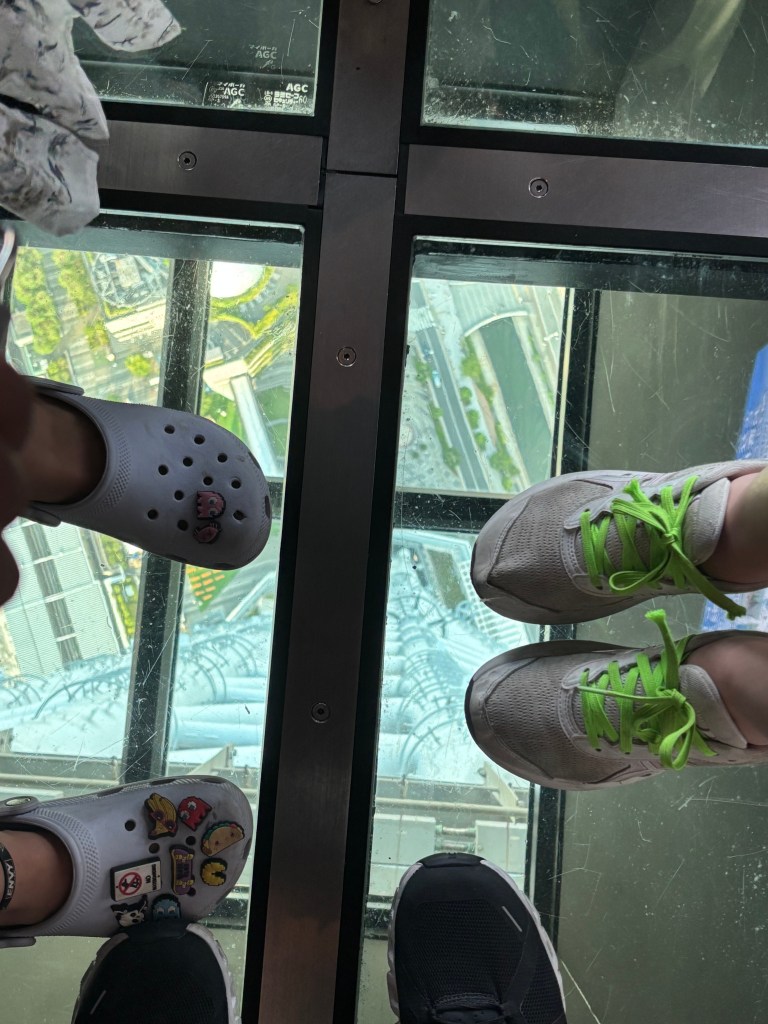 A group of feet standing on a glass floor with a view down to the city below.